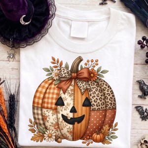 Könnte beinhalten: Ein weißes T-Shirt mit einer großen Grafik eines Patchwork-Kürbisses. Das Kürbisdesign besteht aus Abschnitten mit orangefarbenem Karomuster, Leopardenmuster, Blumenmustern und Punkten, akzentuiert durch eine Leoparden-Schleife und Herbstlaub.