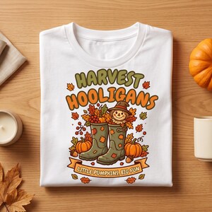 May include: A white t-shirt featuring a graphic of two green rain boots filled with autumn leaves, berries, and a small scarecrow. The text reads "HARVEST HOOLIGANS" and "LITTLE PUMPKINS, BIG FUN" surrounded by orange pumpkins and fall foliage.