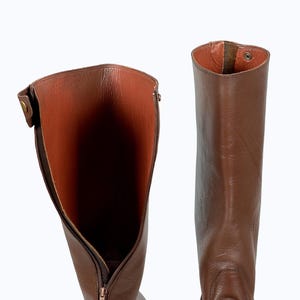 May include: A pair of brown leather riding boots with a zipper on the inside of the boot. The boots are shown with the inside of the boot facing the camera.