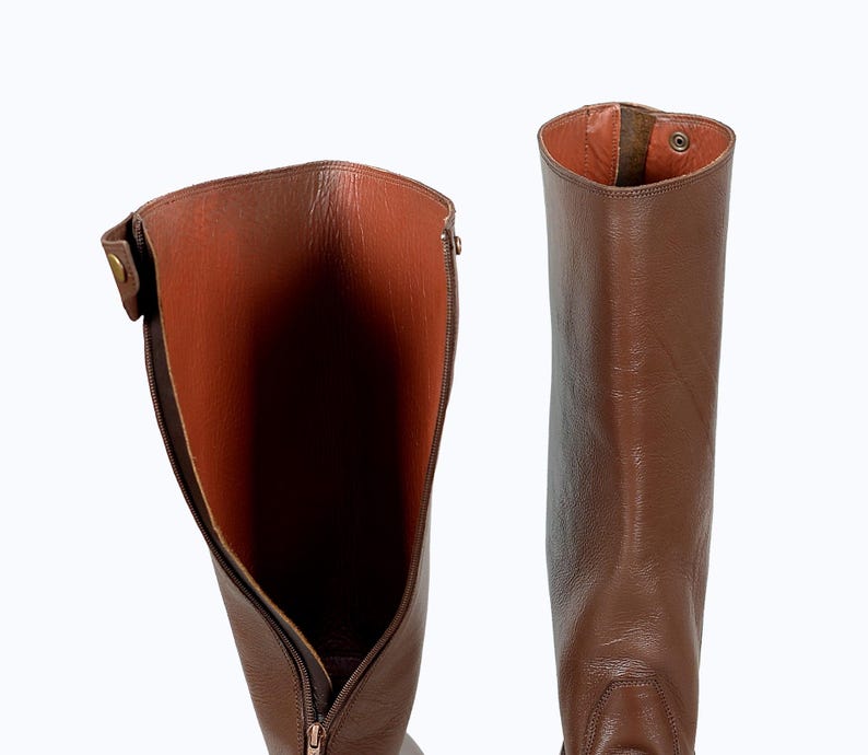 May include: A pair of brown leather riding boots with a zipper on the inside of the boot. The boots are shown with the inside of the boot facing the camera.