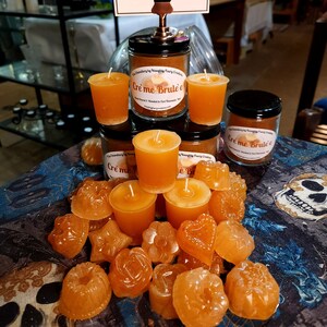 Könnte beinhalten: Eine Sammlung von orangefarbenen Duftkerzen in verschiedenen Formen und Größen, darunter Herzen, Sterne und Schädel. Die Kerzen sind auf einer blau-weißen gemusterten Oberfläche angeordnet. Der Text "Cre'me Brule'e" ist auf dem Etikett eines größeren Kerzenbehälters zu sehen.