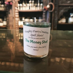May include: A small glass candle with a white label that reads "Naughty Faery Creations Spell Shots! The Money Shot." The candle has a green top and a white wick. The text on the label says "For when you need a quick fix! When you need a face full of money!"