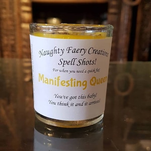 May include: A small yellow candle in a glass jar with a white label that reads "Naughty Faery Creations Spell Shots! For when you need a quick fix! Manifesting Queen You've got this baby! You think it and it arrives!"