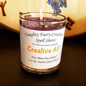 Puede incluir: Una vela morada en un frasco de vidrio con una etiqueta blanca que dice "Naughty Faery Creations Spell Shots! For when you need a quick fix! Creative AF! Your Muse has arrived! Let the creative juices flow!!"
