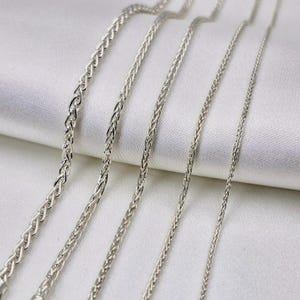 May include: Five silver chains of varying widths and lengths, laid out on a white fabric background.
