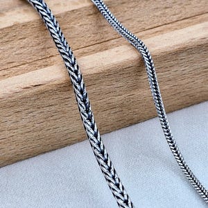 Könnte beinhalten: Zwei silberne Ketten, eine dicker als die andere, liegen auf einer Holzoberfläche. Die Ketten sind beide in geflochtenem Design.