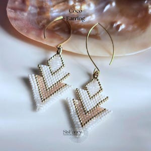 Könnte beinhalten: Coco-Ohrringe mit geometrischem Design. Die Ohrringe sind mit weißen, goldenen und rosafarbenen Perlen versehen. Die handgefertigten Ohrringe haben ein einzigartiges Design. Sie werden auf einer weißen Oberfläche mit einer Muschel im Hintergrund präsentiert.