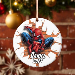 Könnte beinhalten: Runder Weihnachtsschmuck mit Spider-Man, der durch eine Backsteinmauer bricht. Der Superheld ist in einer dynamischen Pose, in einem rot-blauen Anzug. Der Schmuck enthält den Namen "Daniel" und das Jahr "2023".