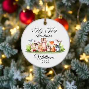 Könnte beinhalten: Weißer Keramik-Weihnachtsschmuck mit dem Text "My First Christmas" und dem Namen "William" mit dem Jahr 2023. Der Schmuck zeigt eine Aquarellillustration von Waldtieren.