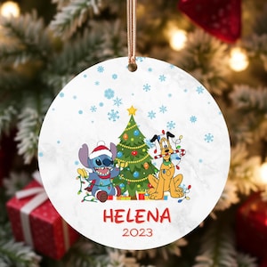 Könnte beinhalten: Runder Weihnachtsschmuck mit Stitch und Pluto neben einem geschmückten Weihnachtsbaum. Der Schmuck trägt den Namen "Helena" und das Jahr "2023" in Rot. Der Hintergrund ist weiß mit blauen Schneeflocken.