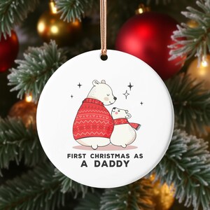 Könnte beinhalten: Runder, weißer Weihnachtsschmuck mit einem Cartoon-Eisbären-Design. Der größere Bär trägt einen roten Pullover, und der kleinere Bär trägt einen roten Schal. Der Text lautet "FIRST CHRISTMAS AS A DADDY".