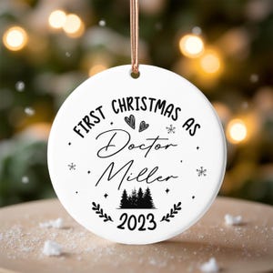 Könnte beinhalten: Weißer Keramik-Weihnachtsschmuck mit dem Text "First Christmas as Doctor Miller 2023". Der Schmuck hat eine goldene Schnur zum Aufhängen und zeigt Herzen, Schneeflocken und eine Waldgrafik.