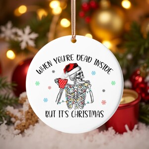 Könnte beinhalten: Runder, weißer Weihnachtsschmuck mit einem Skelett, das einen Weihnachtsmannhut trägt und eine rote Tasse hält. Der Text auf dem Ornament lautet: "WHEN YOU'RE DEAD INSIDE BUT IT'S CHRISTMAS".