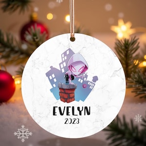 Könnte beinhalten: Runder, weißer Weihnachtsschmuck mit einer Comicfigur in einem Superhelden-Kostüm. Der Schmuck trägt den Namen "EVELYN" und das Jahr "2023". Der Hintergrund zeigt eine Stadtlandschaft und einen Schornstein aus Ziegeln.