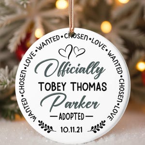 May include: A white ceramic Christmas ornament with the words "Officially Adopted" and the name "Tobey Thomas Parker" in gray script. The ornament is round, with the words "Chosen, Love, Wanted" around the edge. A date, "10.11.21", is at the bottom.