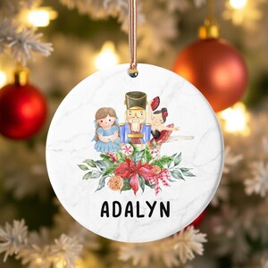 Könnte beinhalten: Weißer Weihnachtsbaumschmuck mit Marmormuster, der eine Aquarellillustration eines Nussknackers, einer Ballerina und eines Mädchens in einem blauen Kleid zeigt. Der Schmuck hat ein roségoldenes Band und den Namen "ADALYN" in schwarzem Text.