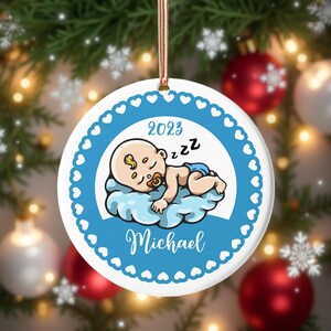 Könnte beinhalten: Weißer Keramik-Weihnachtsschmuck mit einem schlafenden Baby-Design. Der Schmuck hat einen blauen Rand mit Herzformen, den Namen "Michael" und das Jahr "2023". Das Baby schläft auf einer Wolke.