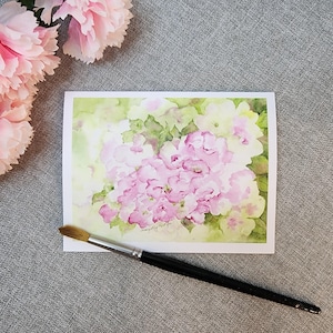 May include: A watercolor painting of pink hydrangeas on a white card, with a green and yellow background. A black-handled paintbrush lies across the card. Pink carnations are in the upper left corner. The painting is signed in the lower right corner.