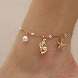 May include: Gold-colored anklet featuring a delicate chain adorned with small pearls. The anklet has three charms: a large pearl, a seashell with a pearl, and a starfish. The anklet is worn on an ankle.
