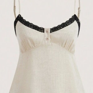 May include: A cream-colored camisole top with adjustable straps and a V-neckline. The top features black lace trim along the neckline and a button-up front. The fabric has a textured appearance.