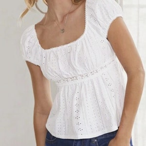 May include: White eyelet top with a square neckline and short, puffed sleeves. The top features a fitted waist and intricate cut-out detailing. A delicate lace trim accents the neckline and waist. The top is made of a lightweight fabric.
