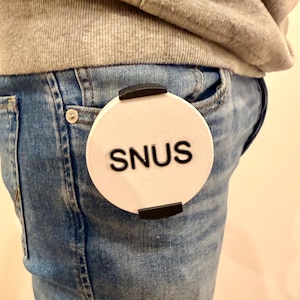 May include: A white, circular product with the word "SNUS" in black lettering is clipped to the pocket of a pair of blue jeans. The product has two black clips on the top and bottom. The person is wearing a grey sweatshirt.