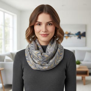 May include: A grey infinity scarf featuring a repeating pattern of small, elongated leaf-like shapes in shades of mustard yellow, navy blue, and white. The scarf is draped in multiple loops around the neck, creating a soft, layered appearance.