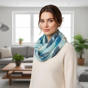May include: An infinity scarf featuring a light blue and dark blue abstract feather pattern. The scarf is worn around the neck over a plain cream-coloured long-sleeved sweater. The fabric displays a soft, layered appearance with varying shades of blue.
