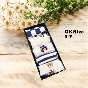 Peut inclure: Une boîte noire contenant cinq paires de chaussettes blanches et bleu marine. Les chaussettes présentent divers motifs, dont un damier, des rayures et des ours en peluche brodés. Le texte "UK Size 3-7" est visible. Des marguerites blanches sont en arrière-plan.