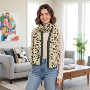 May include: A beige scarf featuring a dark green leaf pattern and a solid dark green border, worn over a beige long-sleeved top and light blue denim jeans. The scarf has a rectangular shape and drapes down the front of the torso.