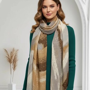Peut inclure: Une écharpe beige, marron et grise avec un motif tacheté. L'écharpe est drapée autour du cou et des épaules. L'écharpe présente un motif de coups de pinceau et de petits points dorés. La personne porte une robe vert foncé.