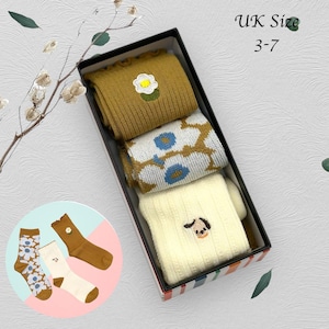 May include: A gift box with three pairs of socks in mustard yellow, cream, and patterned designs. The socks feature embroidered details like a daisy and a dog face. The box is labeled 'UK Size 3-7'. Additional socks are displayed outside the box.