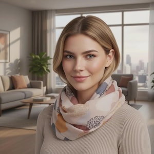 May include: A beige infinity scarf featuring an abstract pattern of muted blue, terracotta, and mustard yellow shapes, accented with small black dashed lines. The scarf is worn around the neck over a ribbed beige long-sleeve top with a crew neckline.