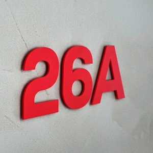 Puede incluir: Número de casa rojo "26A" sobre una pared texturizada gris claro. Los números y la letra están en negrita, en una fuente sans-serif. El color rojo brillante proporciona un contraste llamativo con el fondo neutral.