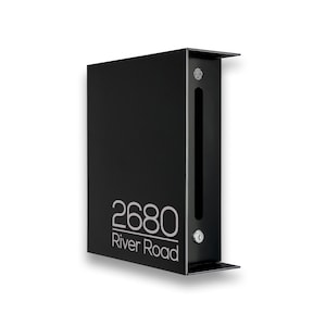 May include: A matte black rectangular wall-mounted mailbox featuring a vertical mail slot and a silver key lock. The front panel displays the white text 2680 River Road in a clean sans-serif font, with a small circular logo at the top right.