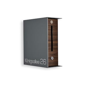 May include: A modern, wall-mounted mailbox with a dark gray metal exterior and a wooden front panel. The text "Königsallee 26" is printed on the front. The mailbox has a slot for mail and a small lock.