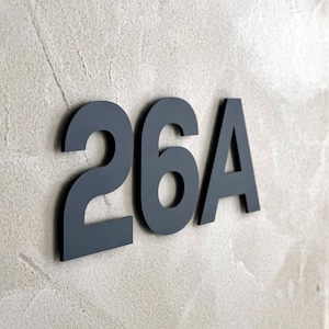 Op de afbeelding: Donkergrijze huisnummers, "26A", zijn gemonteerd op een getextureerde, lichtgrijze muur. De cijfers zijn in een schreefloos lettertype, met een modern, minimalistisch ontwerp. De afbeelding is goed belicht.