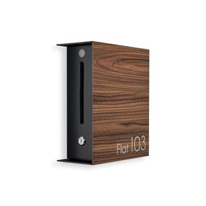 May include: A modern mailbox with a dark wood grain finish and black metal accents. The mailbox has a rectangular shape with the text "Flat 103" in a silver font. The mailbox is mounted on a white wall.