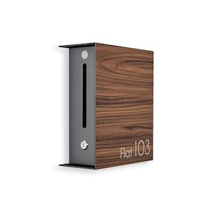 May include: A modern mailbox with a dark wood grain finish and a gray metal front panel. The mailbox has a slot and a keyhole, with the text "Flat 103" in a light color.