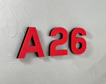 Könnte beinhalten: Rote Holzbuchstaben und -zahlen mit der Aufschrift "A 26" vor einem strukturierten grauen Hintergrund. Die Buchstaben und Zahlen haben eine schwarze Umrandung, die einen 3D-Effekt erzeugt. Das Bild ist gut beleuchtet und die Farben sind lebendig.