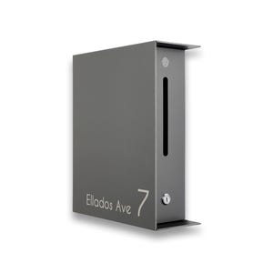 May include: A modern, gray metal mailbox with a sleek, rectangular design. It features a vertical slot, a small lock, and the address "Ellados Ave 7" printed in a minimalist font. The mailbox has a small, angled roof and base.