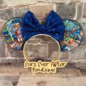 May include: Blue sequined bow and mouse ears headband. The ears feature colorful cartoon graphics and the text "Judy" and "Zootopia". The headband has a gold-colored band and a wooden sign that reads "Ears Ever After Boutique".