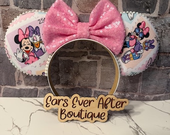 May include: A headband with pink sequined bow and two round ear-shaped decorations. The ear decorations feature Minnie Mouse and Daisy Duck on a postage stamp design. The headband has a wooden sign that reads "Ears Ever After Boutique."