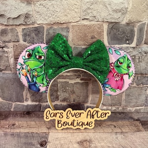May include: A pair of mouse ears with a green sequined bow and two circular ear designs. The ear designs feature a cartoon chameleon character. The headband is gold-colored, and a wooden sign with the text "Ears Ever After Boutique" is at the base.