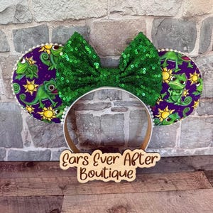 May include: Minnie Mouse ear headband with a green sequin bow and purple ears featuring a cartoon chameleon and sun design. The headband has a white trim and a wooden base. The text "Ears Ever After Boutique" is visible.