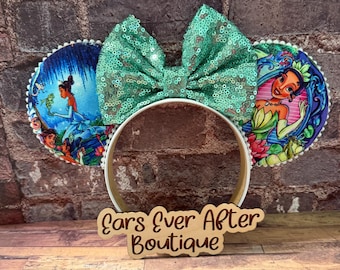 May include: A pair of mouse ears with a teal sequined bow and circular ear designs featuring illustrations of a princess. The headband is white, and the ears are trimmed with white beads. The wooden sign reads "Ears Ever After Boutique."