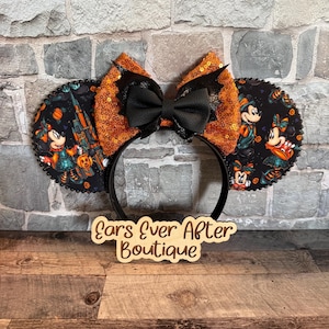 May include: A headband featuring two circular ears with a black fabric background printed with cartoon characters, pumpkins, and a castle. The ears are accented with orange sequins and a central black bow with bat-wing details. A wooden sign reads Ears Ever After Boutique.