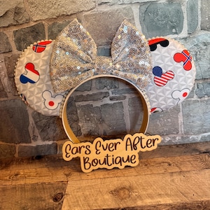 May include: A pair of mouse ears with a silver sequined bow and ear designs featuring flags and Mickey Mouse icons. The headband is white, and a wooden sign below reads "Ears Ever After Boutique."