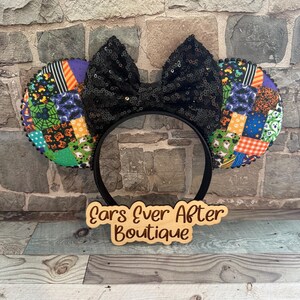 May include: A headband featuring two circular ears covered in a colorful patchwork fabric with Halloween-themed patterns including pumpkins, ghosts, and spiderwebs. A large black sequined bow sits between the ears. A wooden sign reads Ears Ever After Boutique.
