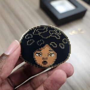 May include: A gold-colored enamel pin featuring a pixelated portrait of a person with brown skin and black hair. The person is wearing a black shirt and has a serious expression.